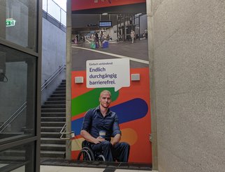 Ein Plakat im Bahnhof auf welchem ein Mensch mit Rollstuhl ist. Mit dem Text: Einfach verbindend: Endlich durchgängig barrierefrei.